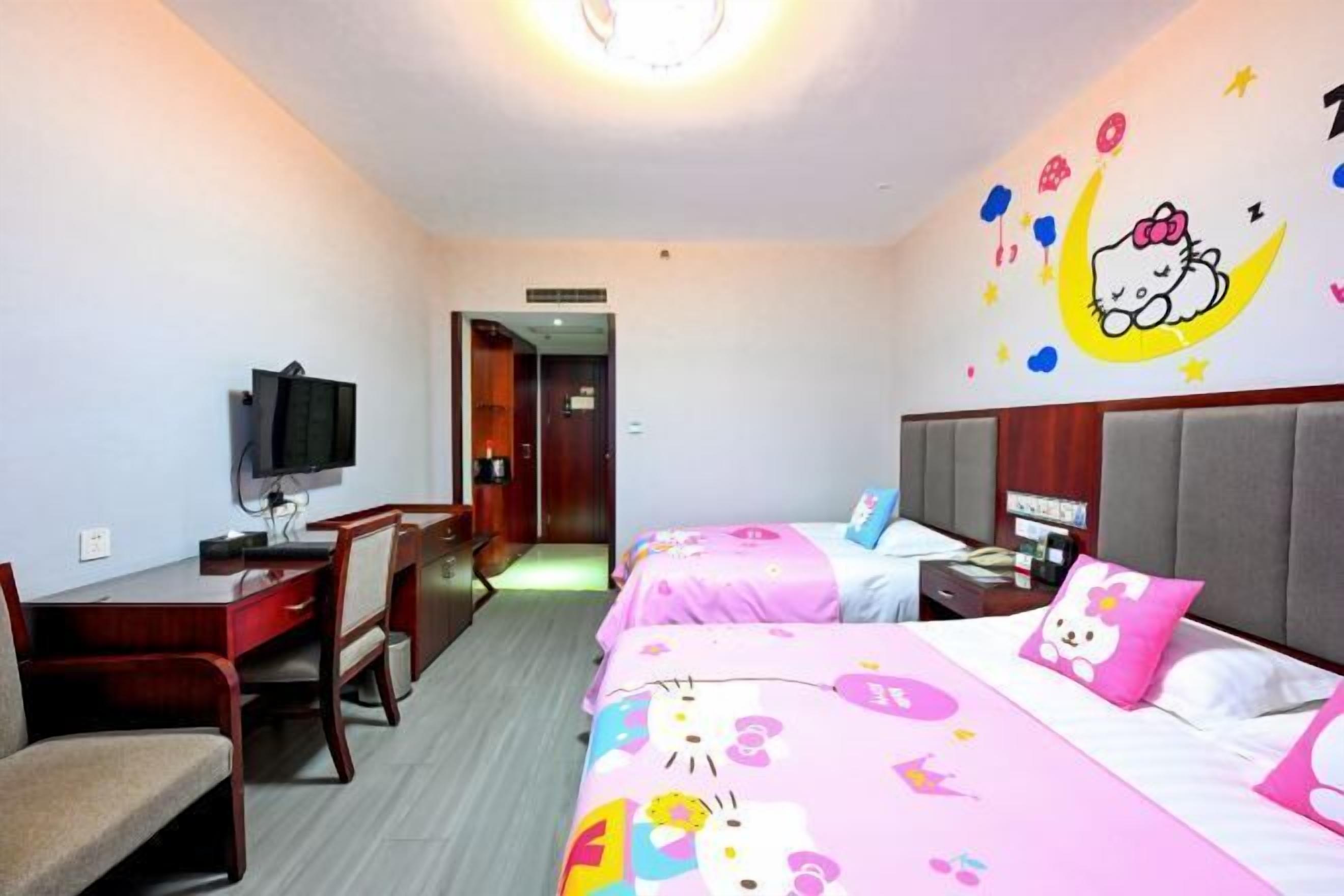 A hotel room with two beds, a desk, a chair, a television, and a wall with a Hello Kitty mural.