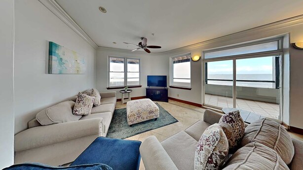 A living room with a sofa, ottoman, and a TV. There are windows and a ceiling fan.