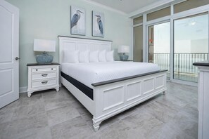 A bedroom with a white bed, a nightstand, a dresser, two wall-mounted lamps, and a balcony with a railing. - Phoenix Gulf Towers 1407 3 Bedroom Condo (Orange Beach)