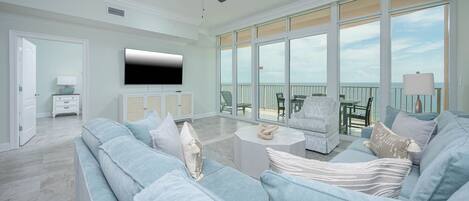 A spacious living room with a large sofa, a television, and a balcony with outdoor furniture.