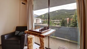 Una habitación con un tablero de ajedrez sobre una mesa de madera, una butaca de cuero negro y un ventanal con vistas a un bosque y edificios.