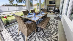 A covered patio with a dining table set for four, surrounded by wicker chairs, a ceiling fan, and a fireplace.
