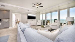 A modern living room with a large sofa, a coffee table, and a TV. There is a balcony with a view of the ocean. - Phoenix Gulf Towers 405 5 Bedroom Condo by RedAwning (Orange Beach)