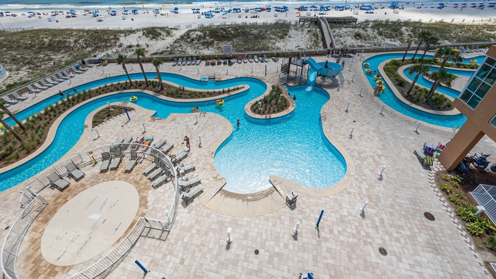 A large outdoor swimming pool with multiple lanes and a slide, surrounded by sun loungers and a sandy beach area.