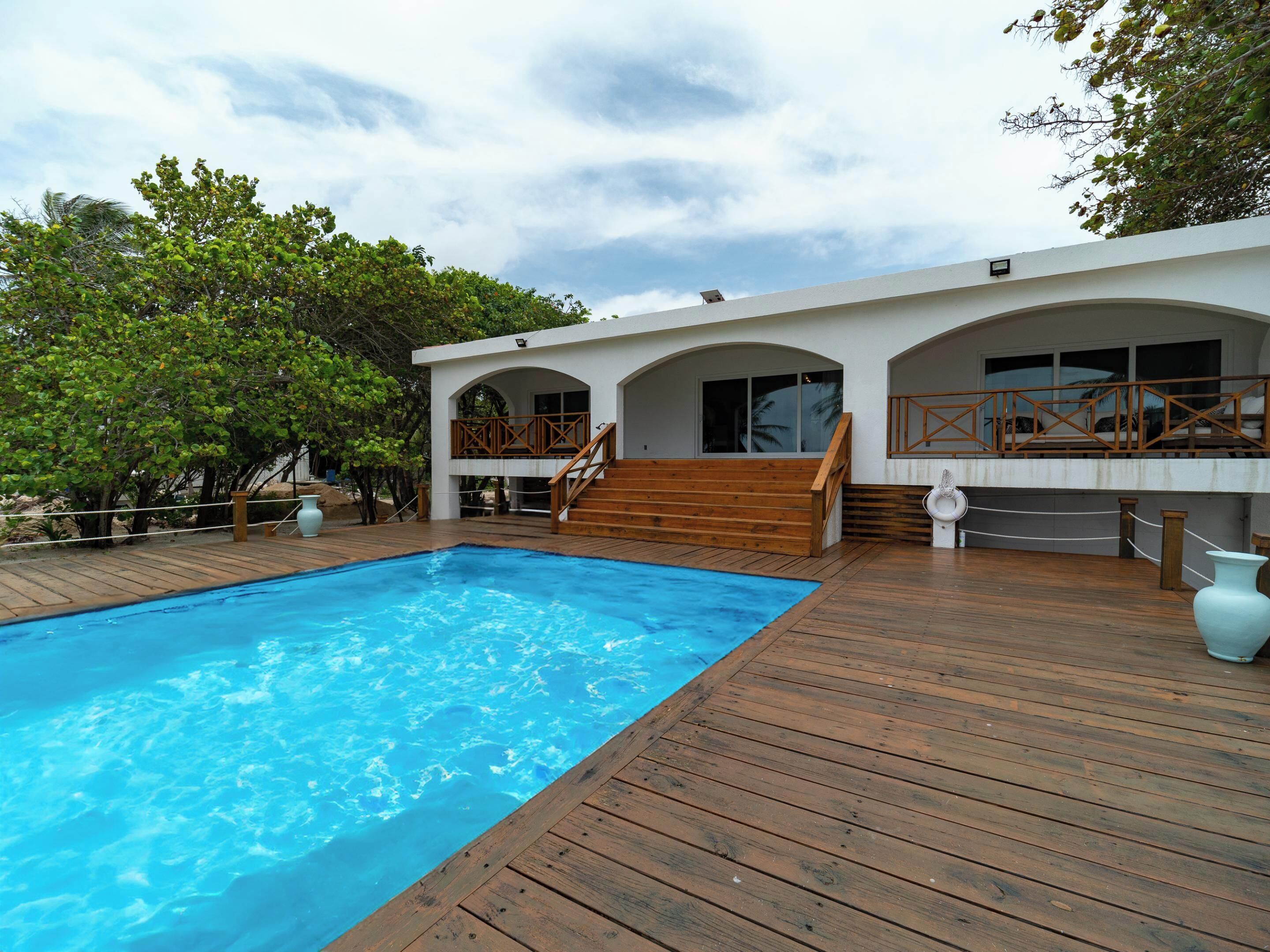 A wooden deck with a swimming pool, a white building with arched windows, and a wooden staircase leading to a balcony.