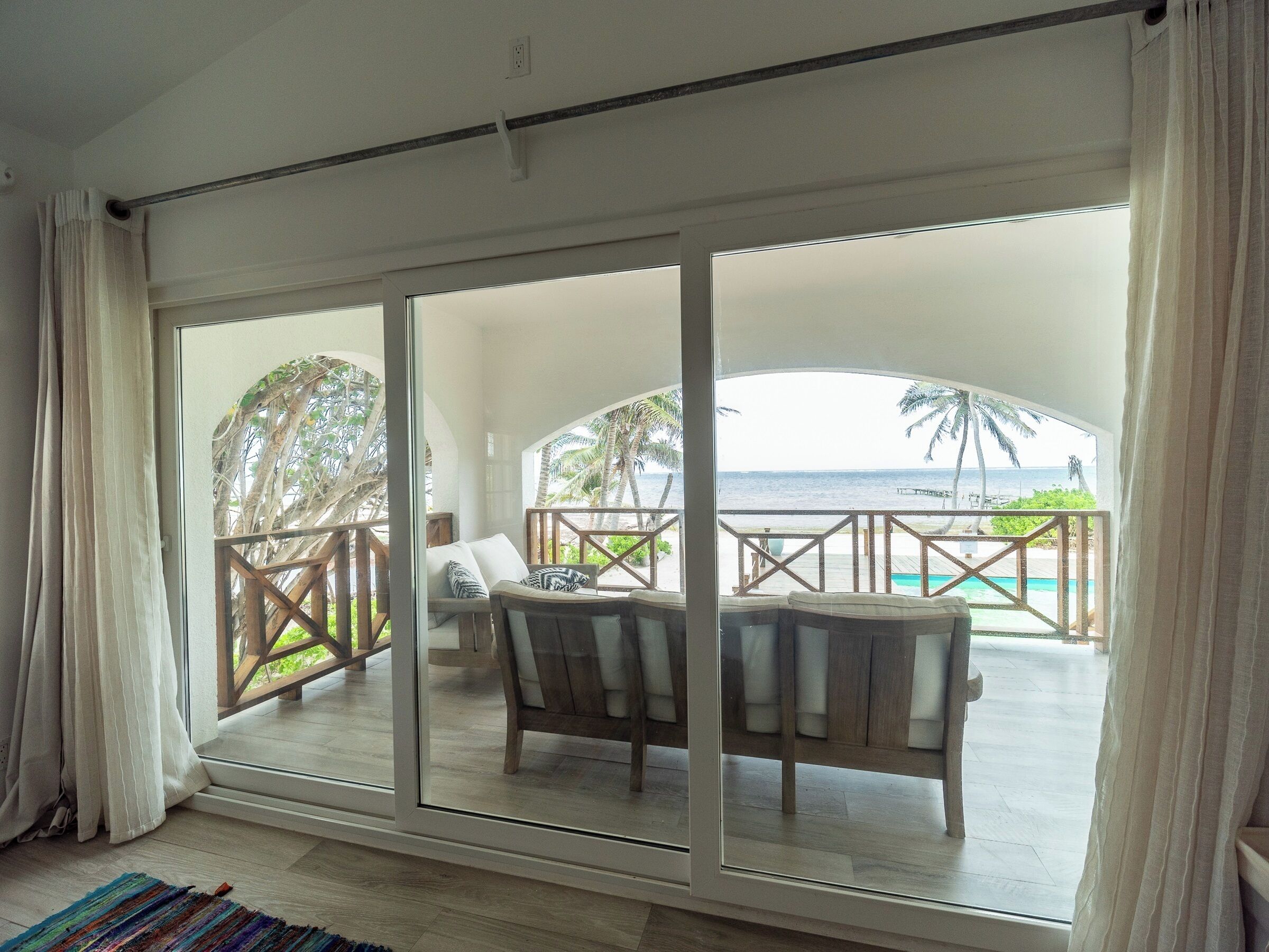 A room with a large sliding glass door leading to a balcony with a view of the ocean and palm trees.