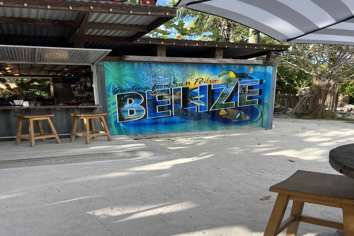 Ein Café direkt am Strand mit einer Wandmalerei, die Belize bewirbt, Sitzgelegenheiten im Freien und sandigem Untergrund.