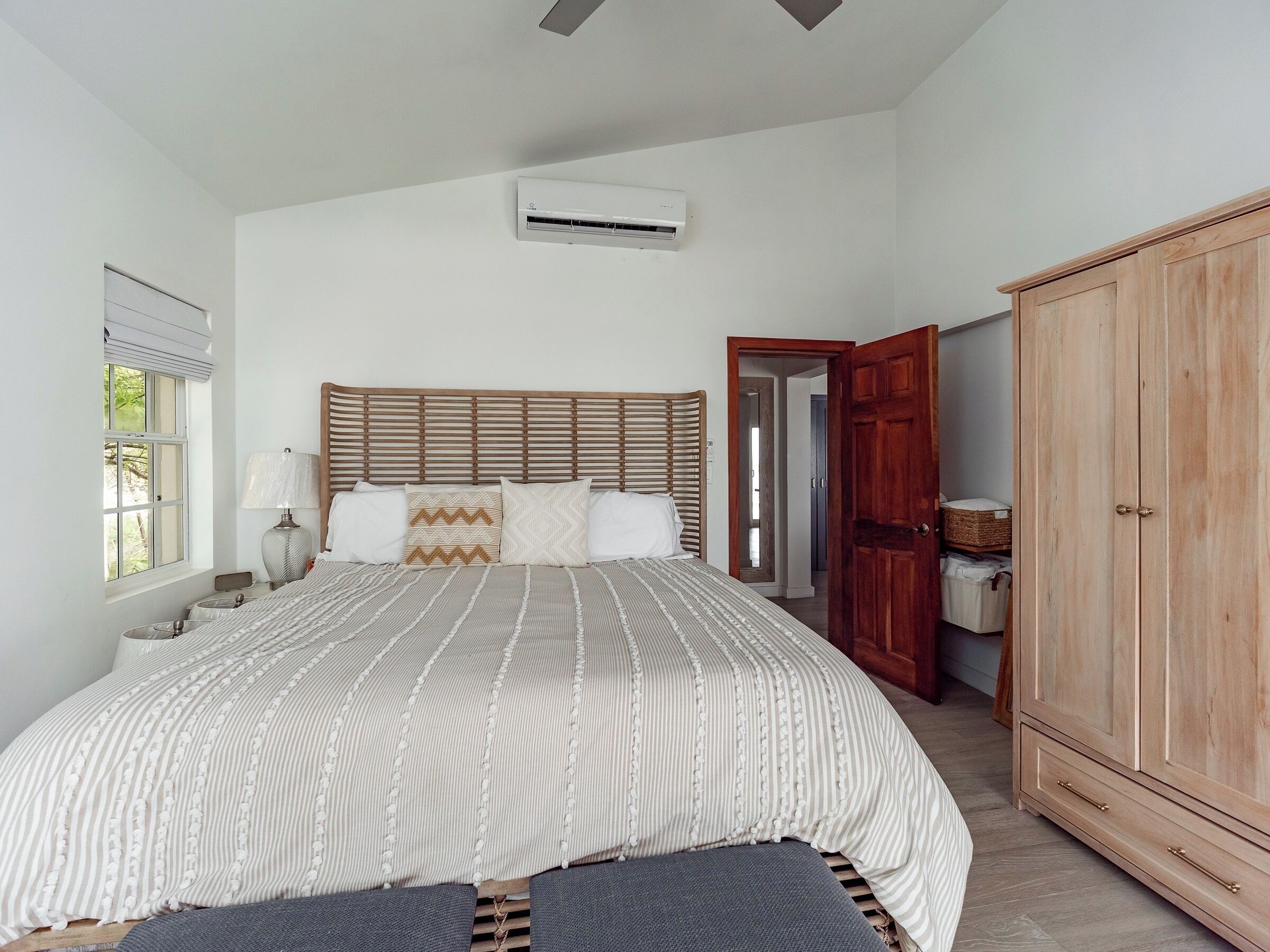 A bedroom with a large bed, a wooden wardrobe, a window with blinds, and an air conditioning unit on the wall.