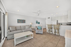 A modern living room with a sofa, a coffee table, a dining area with chairs, a kitchen with cabinets, and a ceiling fan. - Gulf-Front Getaway — Seamist #5 with Bikes & Spectacular Balcony Views (Seacrest)
