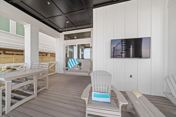 A spacious outdoor deck with a dining area, a seating area with a TV mounted on the wall, and a wooden deck railing.