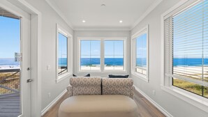 A room with a large window overlooking a beach, a cushioned bench, and a door with a view of the ocean. - "Wait and Sea" New Beachfront Home in PCB with Private Pool and Beach Access! (Panama City Beach)
