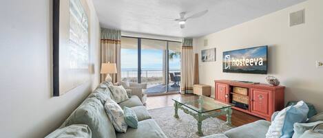 A spacious living room with a large sofa set, a glass coffee table, and a television mounted on the wall.
