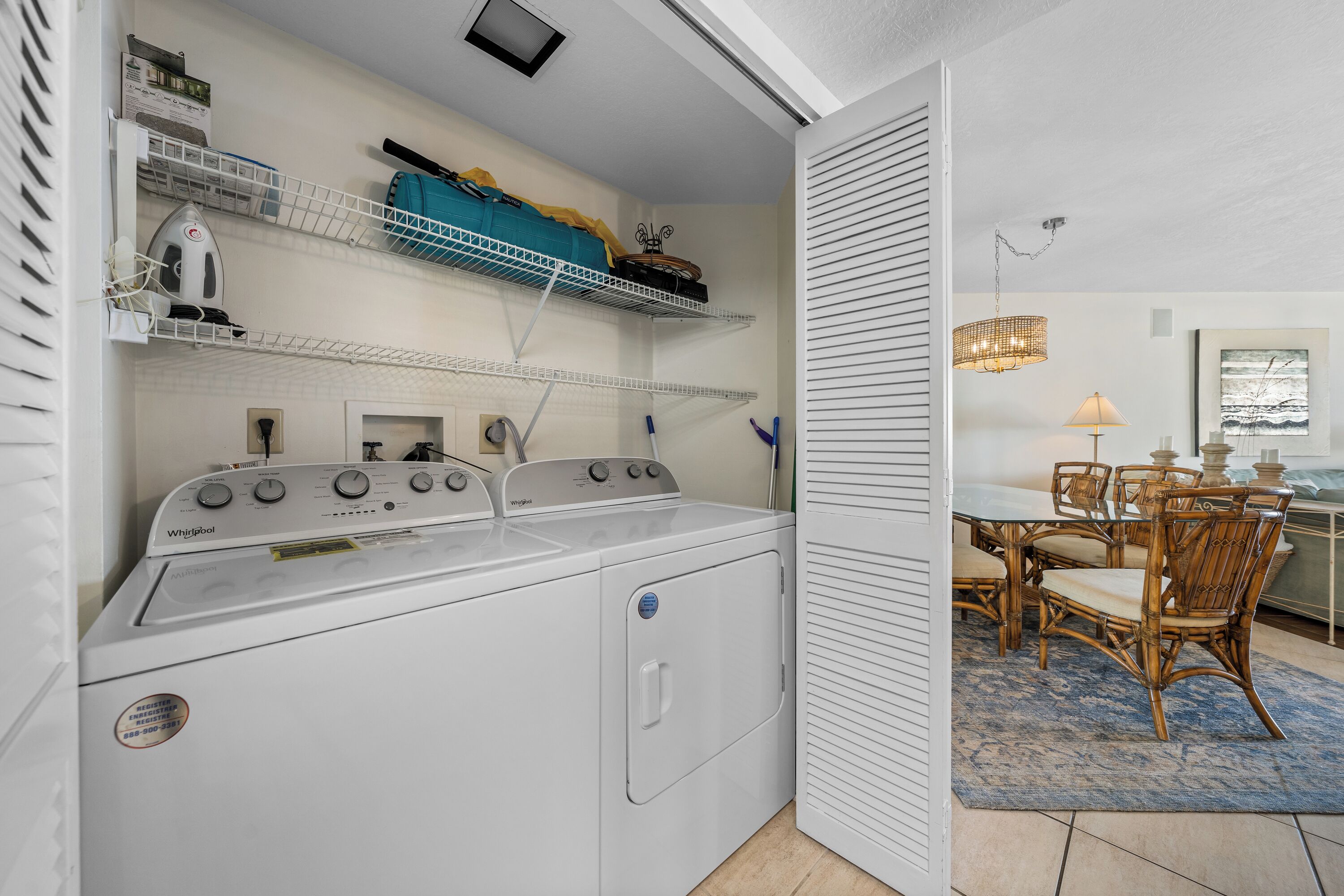 Een wasruimte met een wasmachine en droger, planken met opgevouwen kleding, en een eethoek met rieten stoelen en een tafel.