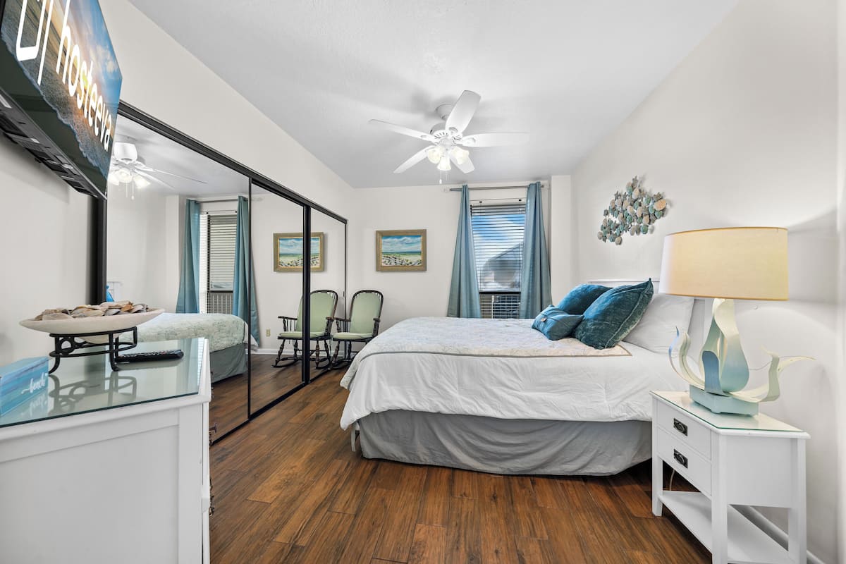 A bedroom with a bed, a nightstand, a mirror, a ceiling fan, and a window with curtains.
