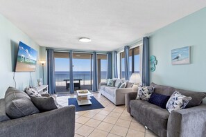 A spacious living room with two sofas, a coffee table, and a TV. Large windows offer a view of the ocean. - White Caps Condos (Orange Beach)