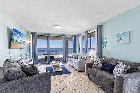 A spacious living room with two sofas, a coffee table, and a TV. Large windows offer a view of the ocean.
