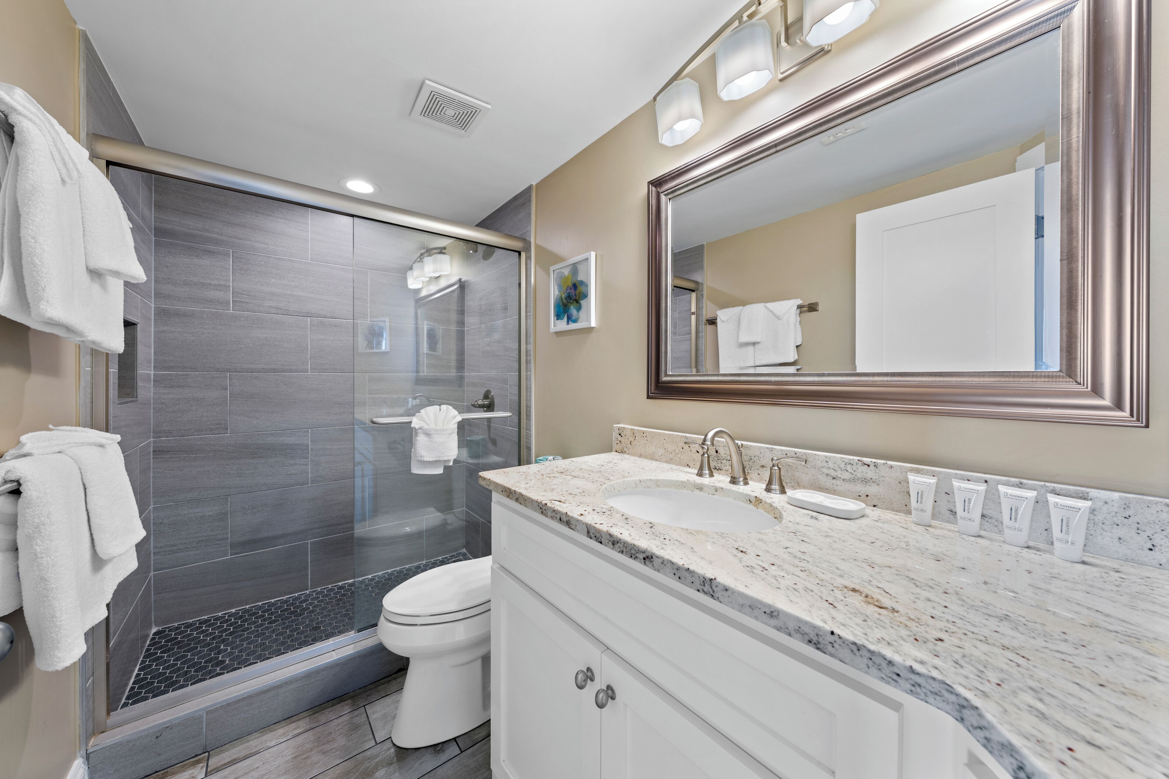 Une salle de bain moderne avec une douche à parois de verre, des toilettes blanches, un grand miroir et un plan de toilette en marbre où sont posés les articles de toilette.