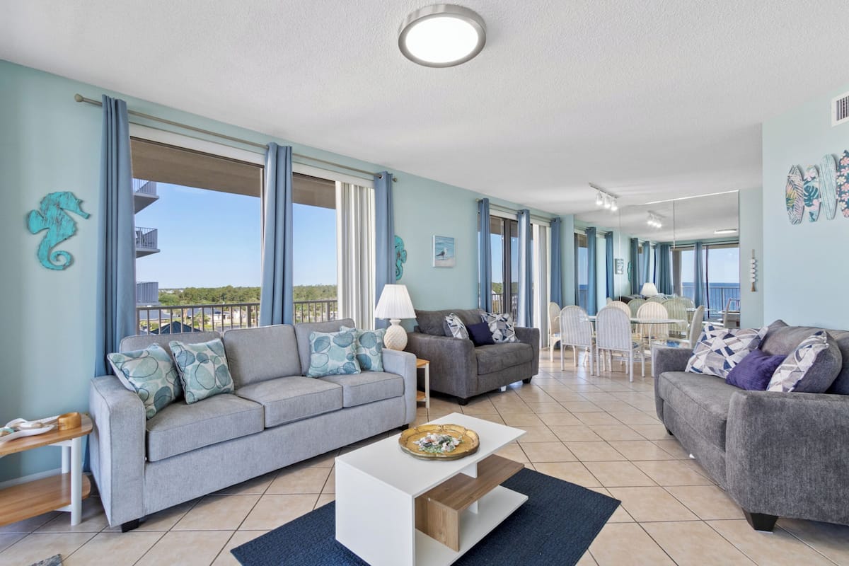 A spacious living area with a grey sofa, a white coffee table, and a dining table with chairs. There is a balcony with a view of the sea.