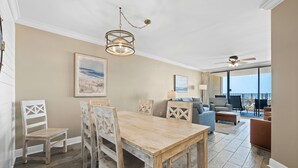 A dining area with a wooden table and chairs, a chandelier, and a view of the ocean through large windows. - White Caps Condos (Orange Beach)