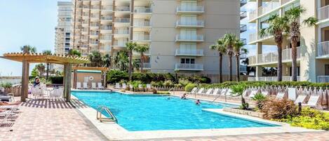 A swimming pool area with lounge chairs and a pergola, surrounded by palm trees and a multi-story building.