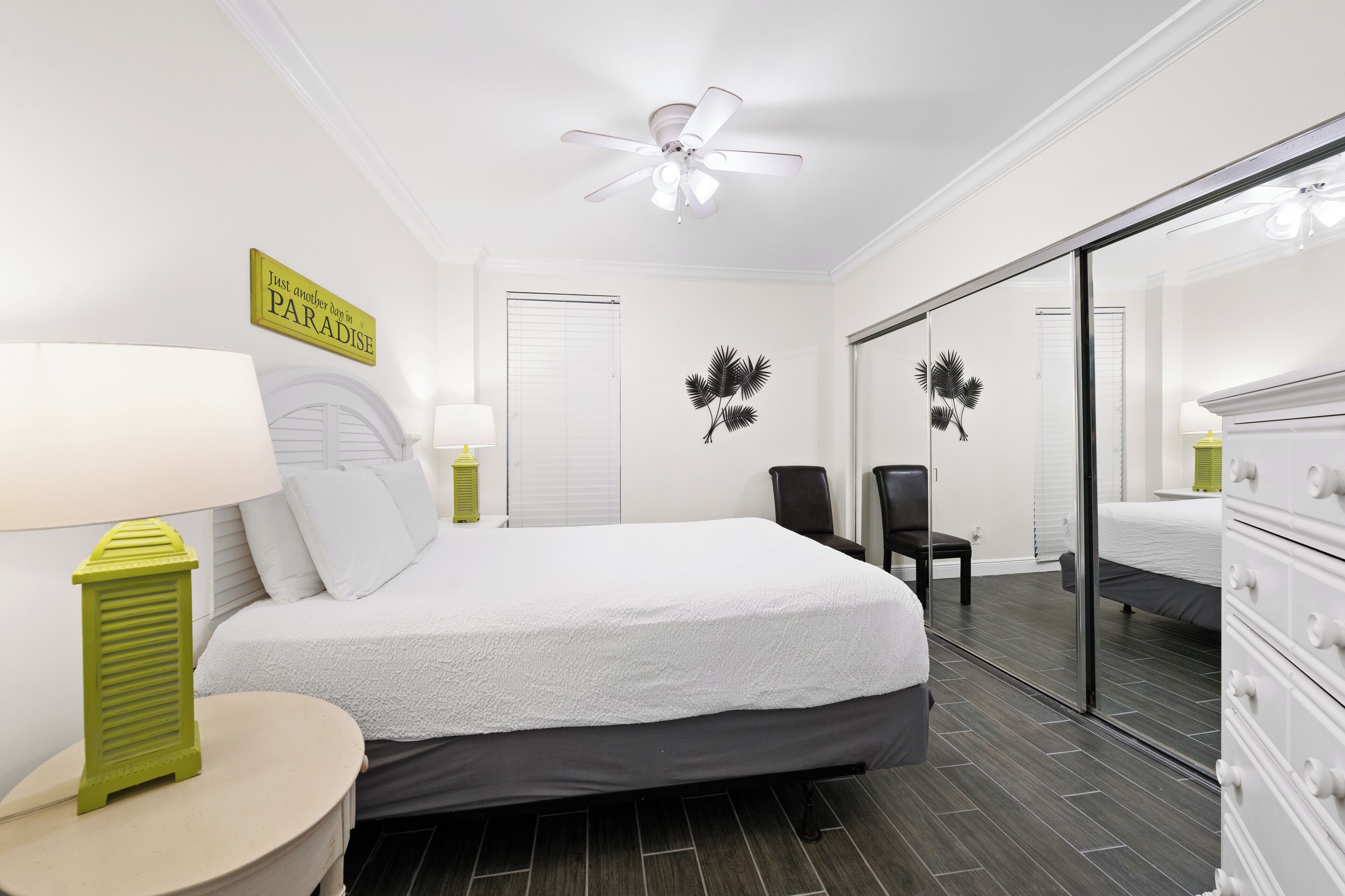 Une chambre à coucher comprenant un lit, une chaise, un miroir, un ventilateur de plafond et un mur orné d’un panneau décoratif.