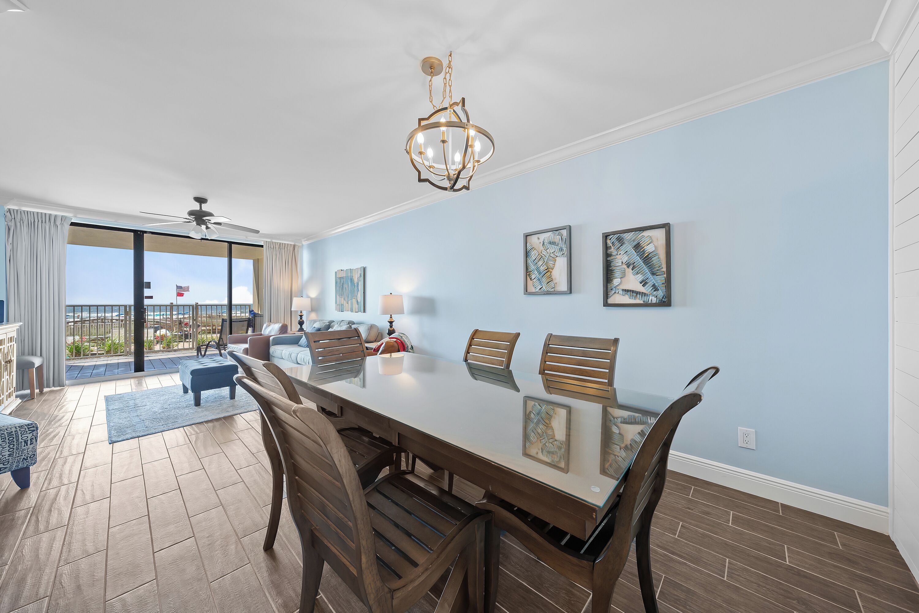 A spacious dining area with a large table, wooden chairs, and a chandelier. There are two framed artworks on the wall and a ceiling fan.