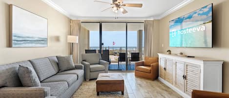 A modern living room with a grey sofa, a brown leather chair, a ceiling fan, a television, and a view of the ocean through large sliding doors.