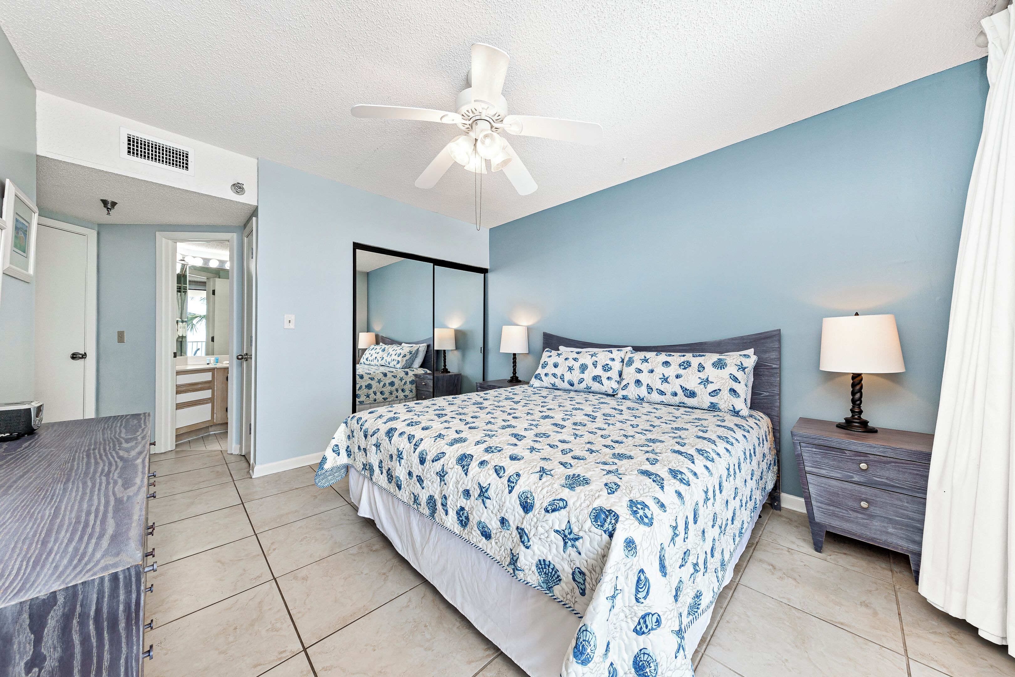 A bedroom with a bed, bedside tables, a ceiling fan, a mirror, and a bathroom door.