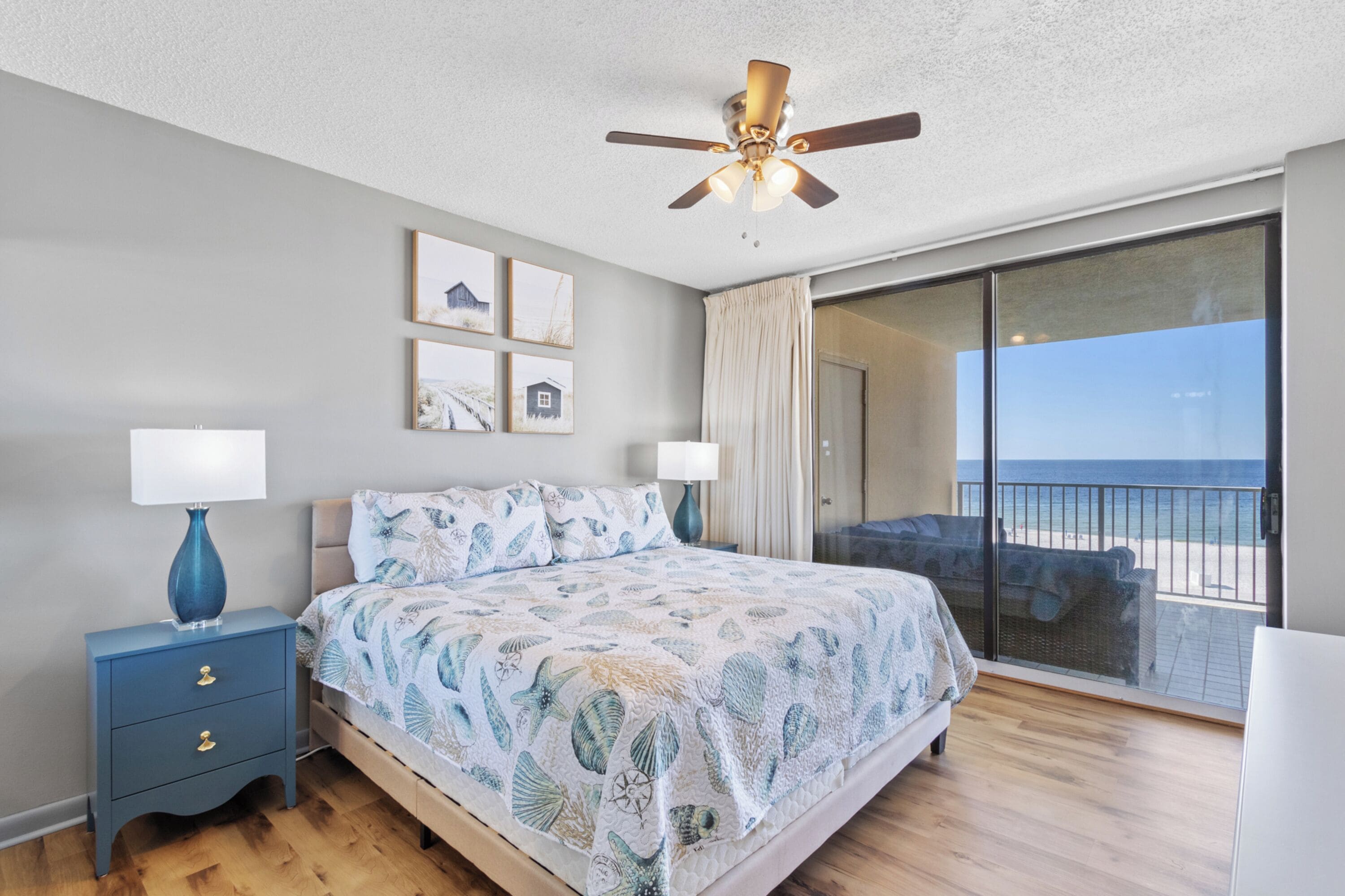 A bedroom with a bed, nightstands, a ceiling fan, and a balcony with a view of the beach.
