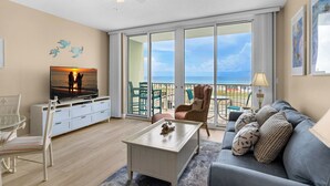 Ein großzügiges Wohnzimmer mit großem Fernseher, bequemen Sitzmöglichkeiten, einem Balkon mit Meerblick und einem Deckenventilator.