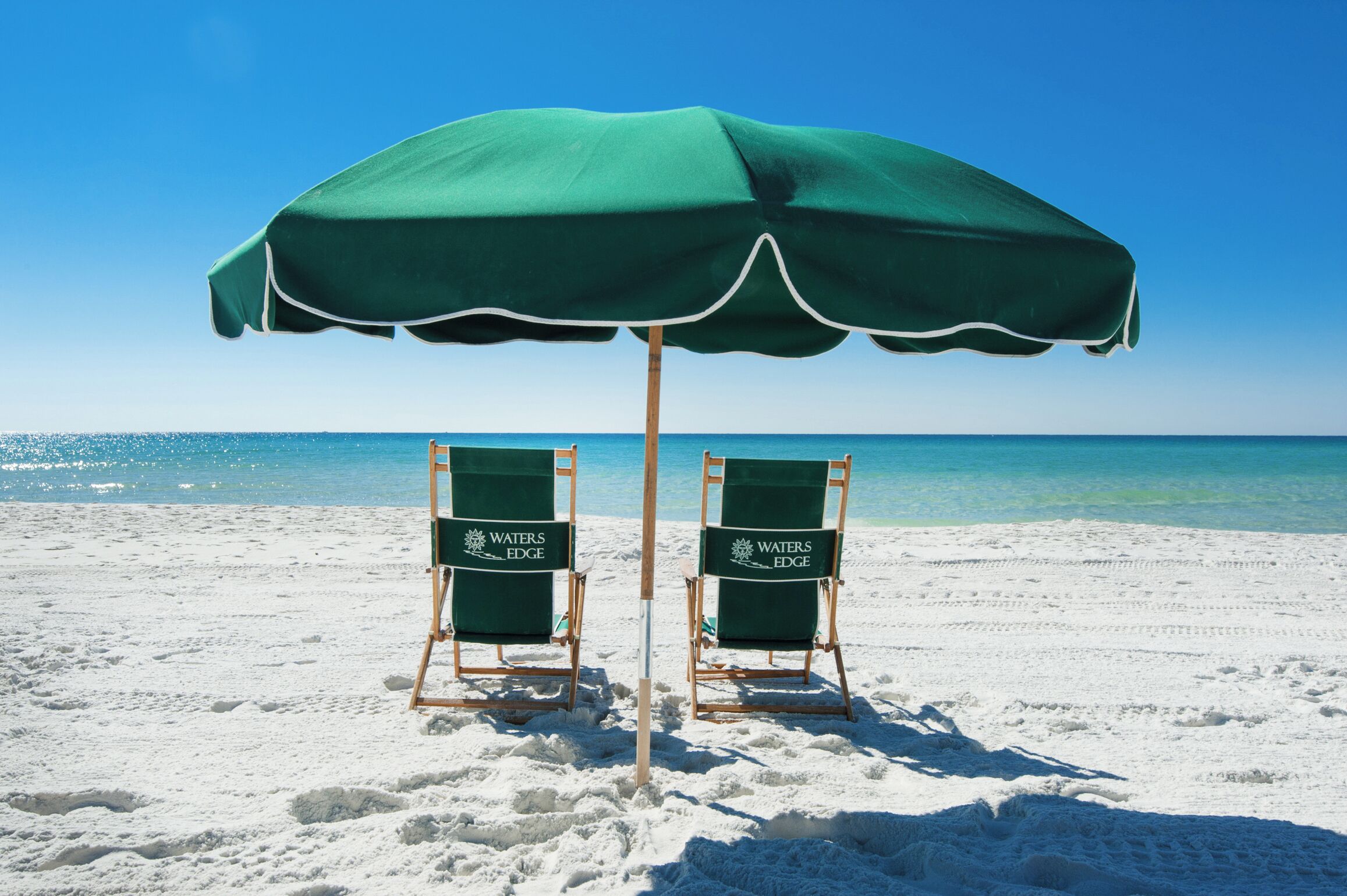 Two beach chairs under a green umbrella on a sandy beach with clear blue water in the background.