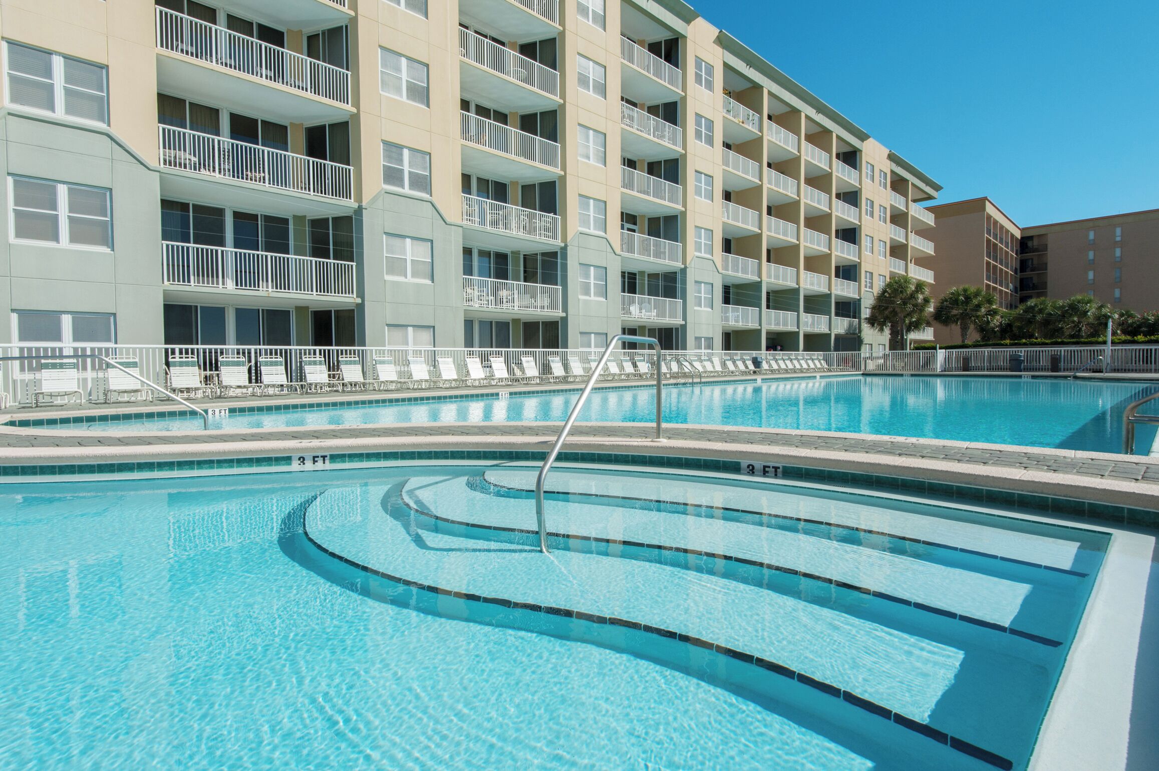 A swimming pool with multiple lanes, surrounded by a building with balconies and a clear blue sky.