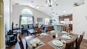 Un vaste espace dédié à la salle à manger, avec une grande table en bois dressée pour quatre personnes, une cuisine à l’arrière-plan et un salon doté d’un canapé et d’un ventilateur de plafond.