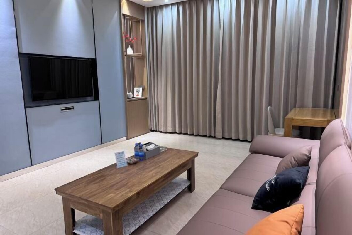 A modern living room with a wooden coffee table, a sofa, a TV mounted on a cabinet, and a large window with curtains.
