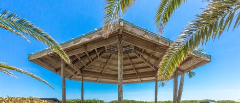 A wooden pavilion with a roof, surrounded by palm trees and picnic tables.