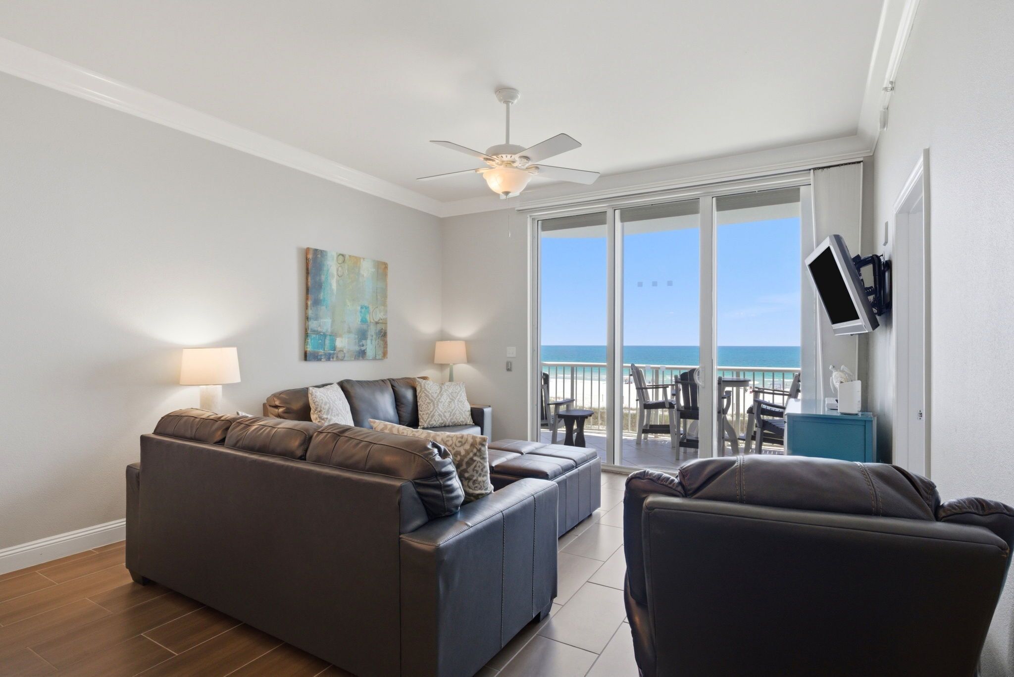 Ein Wohnzimmer mit einer Couch, einem Sessel, einem Deckenventilator, einem an der Wand montierten Fernseher und einem Balkon mit Meerblick.