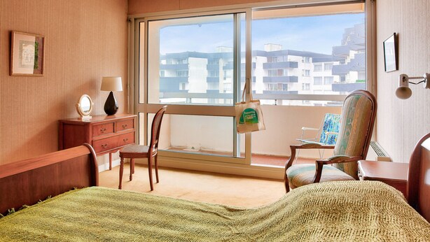 Una habitación de hotel con un ventanal grande, una cama, un aparador, una silla y una mesita.