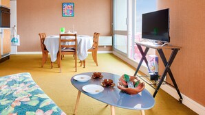 Una habitación de hotel compacta con zona de comedor, un televisor sobre un mueble bajo y un ventanal grande.
