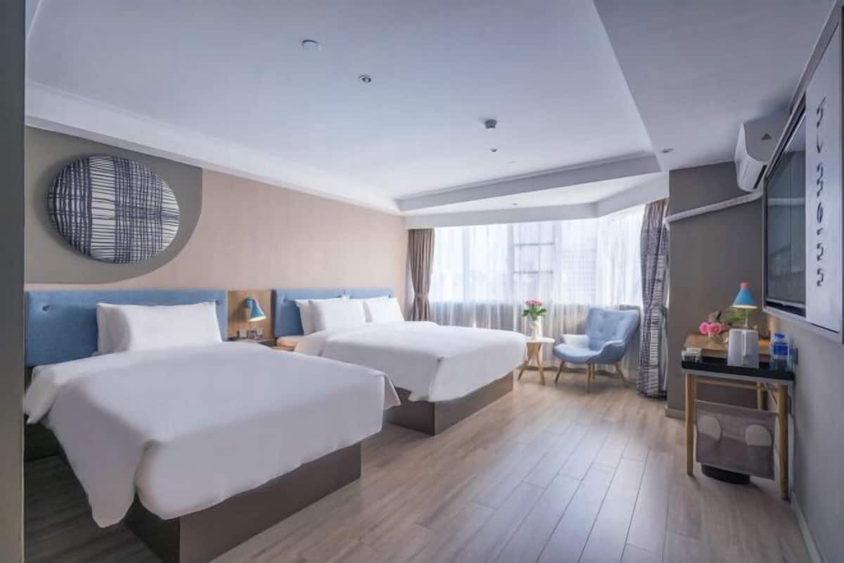A hotel room with two beds, a round wall decoration, a TV, a small table with a lamp, and a view of the outside through large windows.
