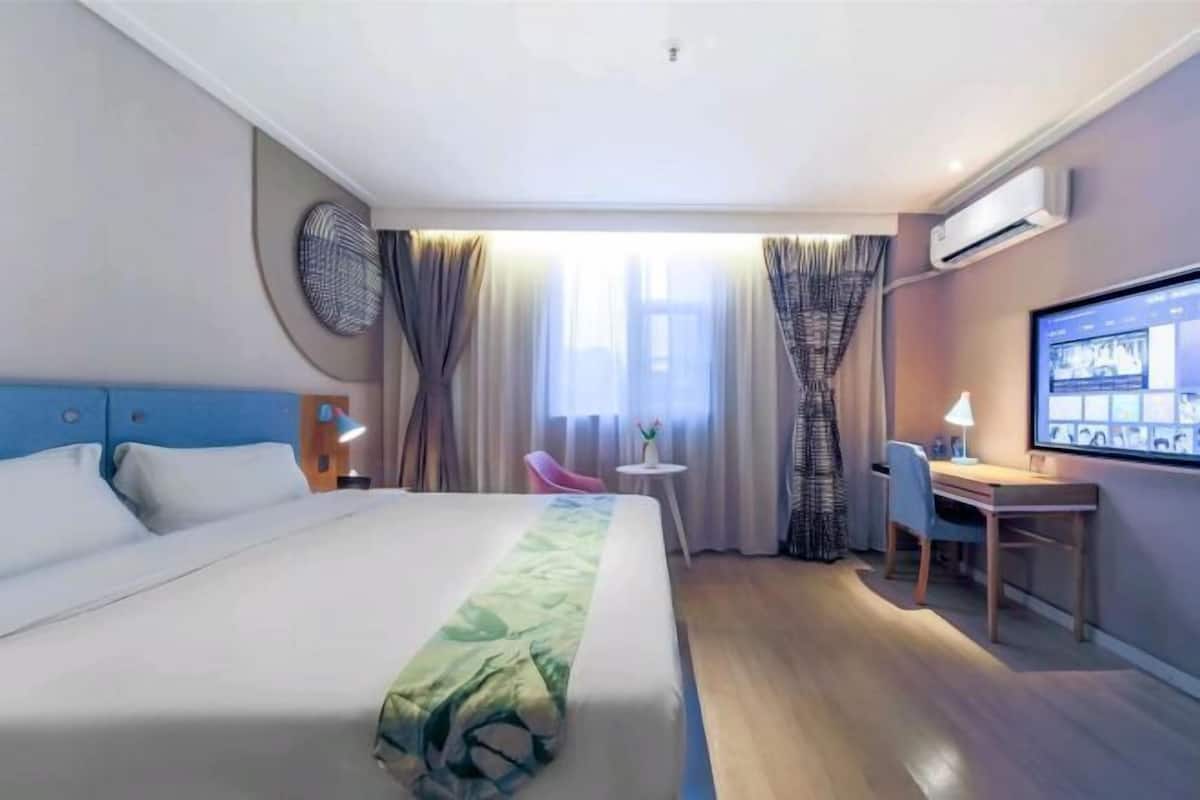 A hotel room with a large bed, a desk, a chair, a TV, and an air conditioning unit.