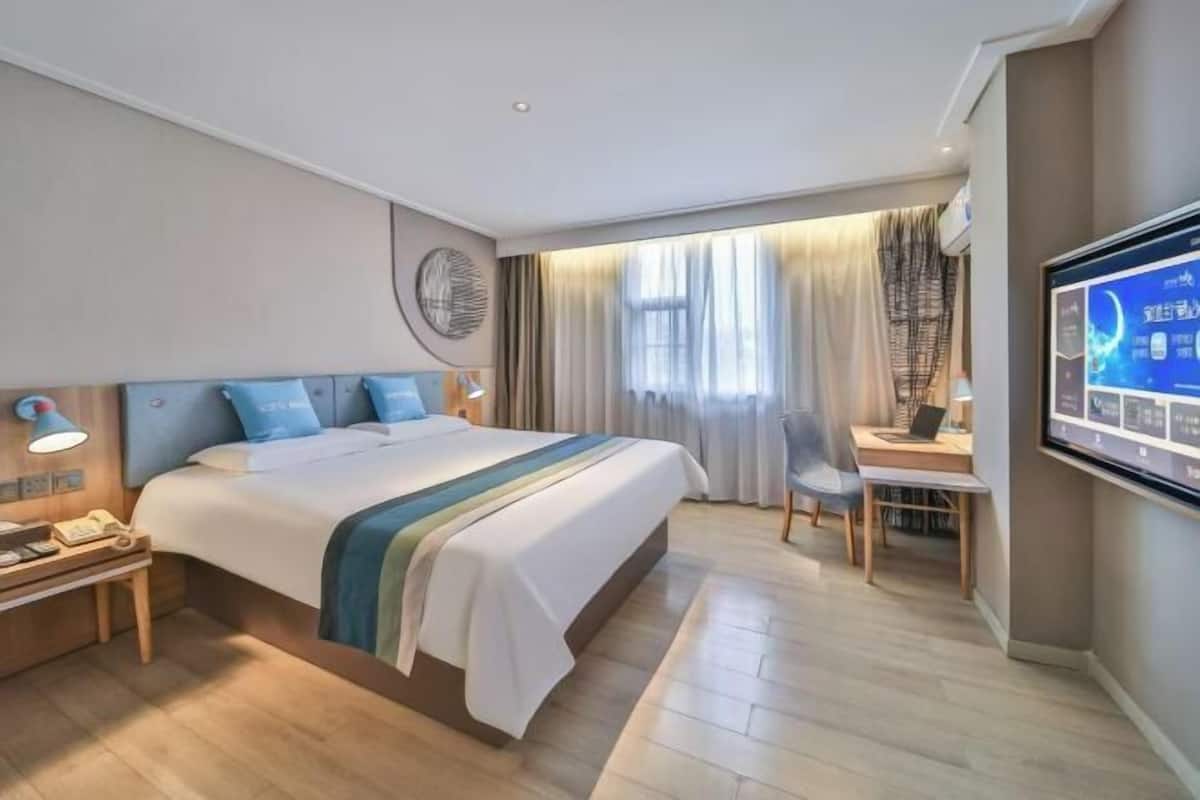 A hotel room with a bed, desk, chair, TV, and a wall-mounted control panel.