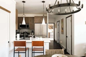 A modern kitchen with stainless steel appliances, a central island, pendant lighting, and a dining area with two chairs. - New Luxe Condo | Hot Tub | Minutes to WP! | Fraser (Fraser)