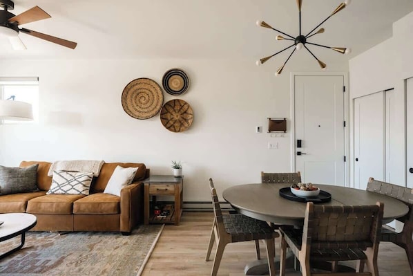 A modern living room with a brown sofa, a dining table with chairs, a ceiling fan, and wall-mounted decor.