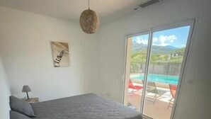 Ein Schlafzimmer mit Bett, Nachttisch mit Lampe, Wandbild und Blick auf einen Swimmingpool durch eine Schiebeglastür.