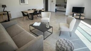 Ein großzügiger Wohnbereich mit einer Couch, einem Couchtisch und einem gemusterten Hocker. Ein Esstisch mit Stühlen und ein Fernseher sind im Hintergrund zu sehen.