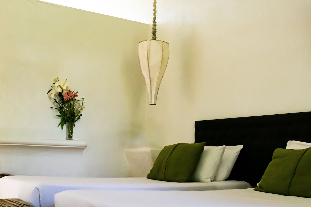 A hotel room with two beds, a small table, a hanging light fixture, and a wall-mounted shelf with flowers.