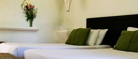 A hotel room with two beds, a small table, a hanging light fixture, and a wall-mounted shelf with flowers.