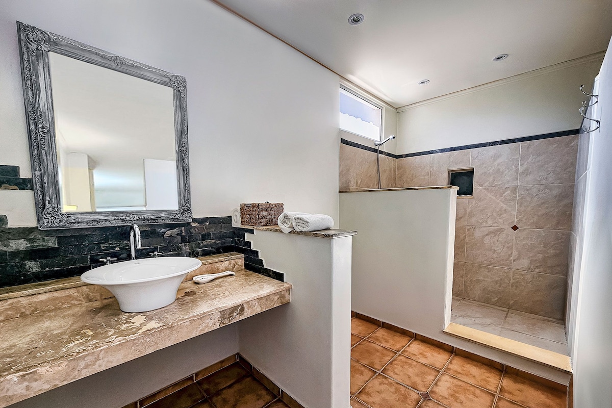 Un baño con lavamanos blanco, un espejo grande, ducha y piso de baldosas.