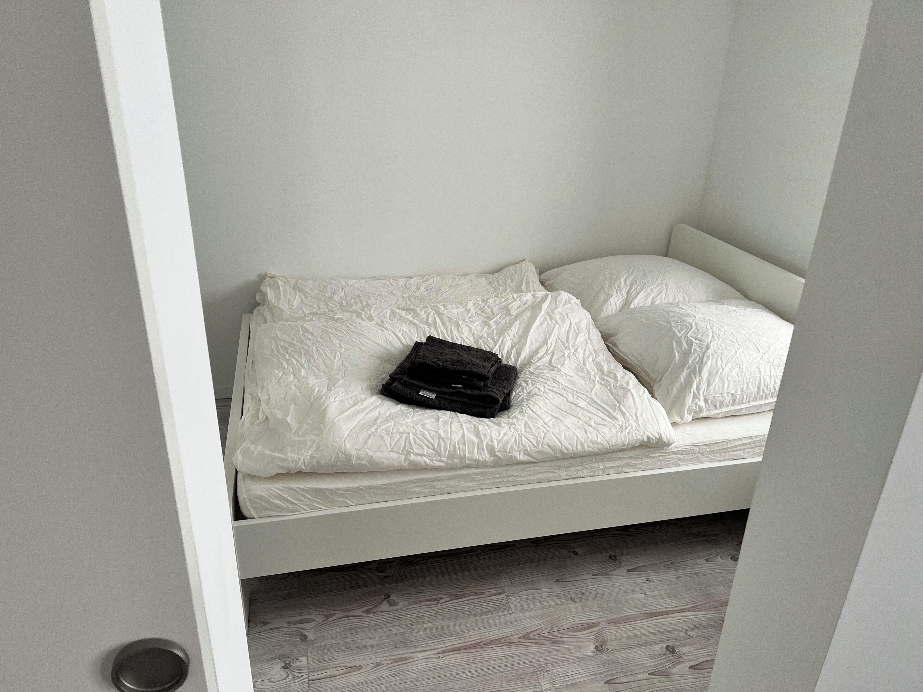 Ein weißes Bett mit zwei Kissen und einer zusammengelegten Decke in einem Zimmer mit Holzboden.