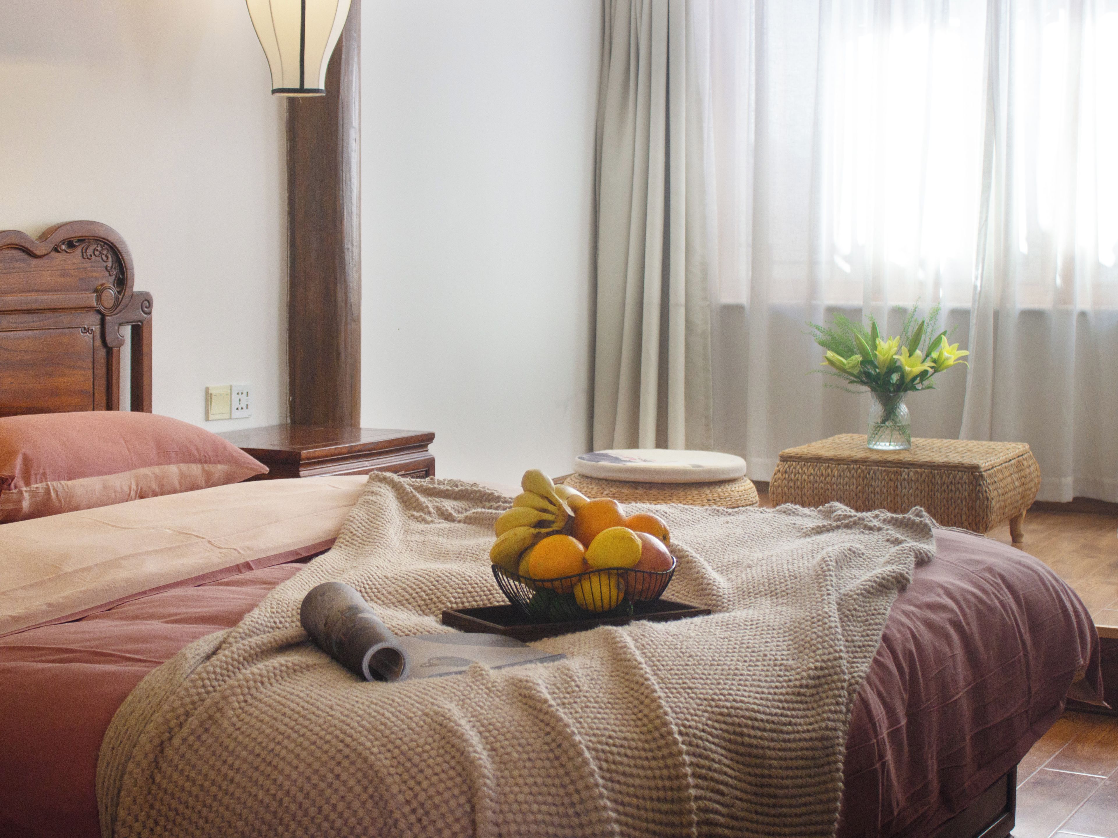Ein ordentlich bezogenes Bett mit einer gestrickten Decke, ein Nachttisch mit Obstschale und eine Lampe.